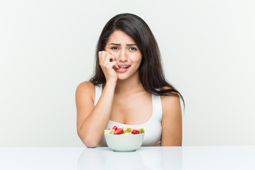 Young hispanic woman eating a fruit bowl biting fingernails, nervous and very anxious.