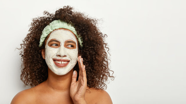 Satisfied Afro American lady has day spa, wears white fasial mask, touches cheek, has natural beauty, curly luminous hair, focused aside, wears headband, stands shirtless indoor. Copy space for advert
