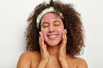Headshot of pleased relieved woman massages face with sea salt, smiles happily, keeps eyes shut...