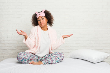 Young african american woman in the bed wearing pijama doubting and shrugging shoulders in...