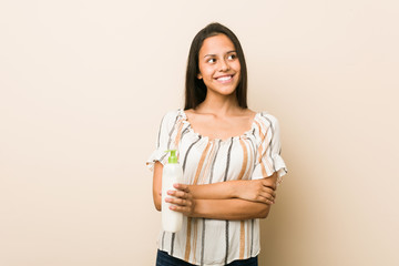 Young hispanic woman holding a cream bottle smiling confident with crossed arms.