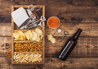 Glass and bottle of craft lager beer and opener with box of snacks on wooden background. Pretzel...