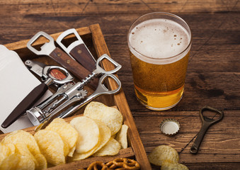 Glass of craft lager beer and opener with box of snacks on wooden background. Pretzel and crisps and salty potato sticks in vintage wooden box with openers and beer mats. Macro