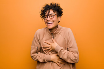 Young mixed african american woman against a brown background laughs happily and has fun keeping hands on stomach.