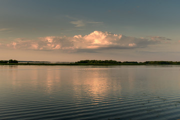 Golden sunset clouds are reflected in the water of a wide quiet river against the background of islands covered with green trees.