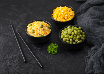 Black bowls with boiled organic basmati vegetable rice, yellow corn and peas with black chopsticks on black stone background with linen cloth. Top view