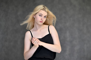 Portrait of a cute blonde student girl, a young woman in a black dress with beautiful curly hair on a gray background in different poses. Shows hands to the sides. Beauty, brightness, emotions.