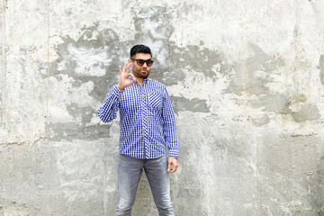 Obraz na płótnie Canvas Its Ok. Portrait of satisfied handsome bearded young man in checkered blue shirt and sunglasses standing against concrete gray wall, showing Ok sign and looking at camera with satisfied face.