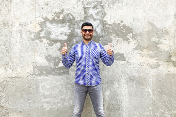 I like it. Portrait of happy satisfied handsome bearded young man in checkered blue shirt and sunglasses standing against concrete gray wall. thumbs up and looking at camera with toothy smile.