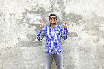 Portrait of hopeful handsome bearded young man in checkered blue shirt and sunglasses standing against concrete gray wall. crossed finger, clenching teeth and hope or wish to ne lucky or winner.