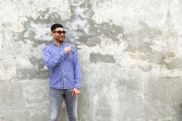 Obraz na płótnie Canvas Portrait of happy handsome bearded young man in checkered blue shirt and sunglasses standing against concrete gray wall. pointing at background empty copyspace and looking at camera with toothy smile.