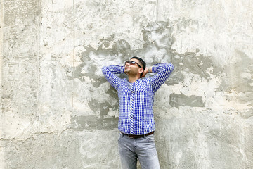 Obraz na płótnie Canvas Portrait of happy handsome bearded young man in checkered blue shirt and sunglasses standing against concrete gray wall. holding his head and looking away with pondering dreaming face and smiling.