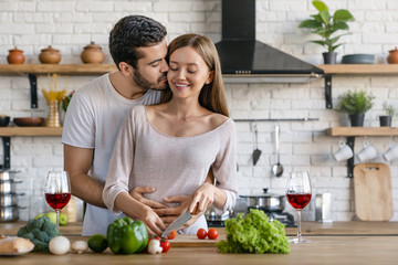 Beautiful cheerful young couple cooking dinner while man kissing his girlfriend standing in the...