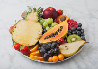 Fresh raw organic summer berries and exotic fruits in white plate on marblebackground. Pineapple, papaya, grapes, nectarine, orange, apricot, kiwi, pear, lychees, cherry and physalis.