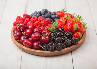 Fresh organic summer berries mix in round wooden tray on light wooden table background. Raspberries, strawberries, blueberries, blackberries and cherries.