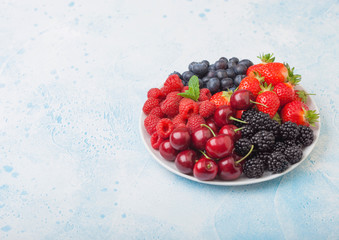 Fresh organic summer berries mix in white plate on blue kitchen table background. Raspberries, strawberries, blueberries, blackberries and cherries. Space for text