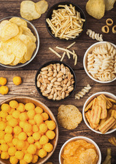 All classic potato snacks with peanuts, popcorn and onion rings and salted pretzels in bowl plates on wooden background. Twirls with sticks and potato chips and crisps with nachos and cheese balls.