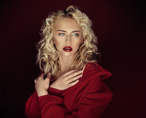 Beautiful portrait seductive woman with light blond hair,blue eyes and red lips posing in red coat and on dark red background. Fashion, glamour, concept