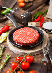 Fresh raw minced homemade farmers grill beef burgers on round chopping board and frying pan with spices and herbs and meat hatchet on wooden board. Oil, garlic and tomatoes.