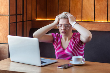 Portrait of illness young adult woman with short blonde hair in pink t-shirt and eyeglasses sitting in cafe and holding head with hands have a high pain, headache and migraine. Indoor, medical concept