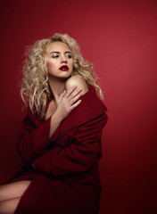 Naklejka premium Beautiful portrait seductive woman with light blond hair,blue eyes and red lips posing in red coat and on dark red background. Fashion, glamour, concept
