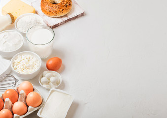 Fresh dairy products on white table background. Glass of milk, bowl of sour cream and cottage cheese and eggs. Fresh baked bagel. Steel whisk. Top view.Space for text