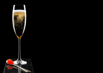 Elegant glass of yellow champagne with red caviar on golden spoon on marble board on black background. Space for text