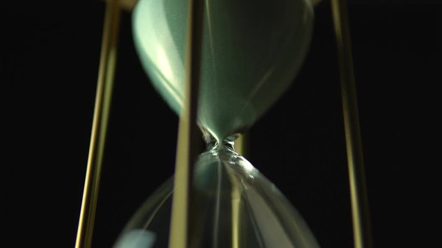 Slow motion rotation of sand timer clock with sand flowing from through hour glass. View from below hourglass close up macro shot. Black background.