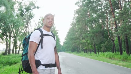 Handsome African man hitchhiking on countryside road with backpack. Attractive cheerful male hitchhiker catching a ride during his trip across country