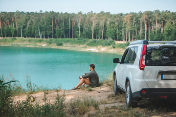 Obraz na płótnie Canvas man sitting near white suv car at the edge looking at lake with blue water