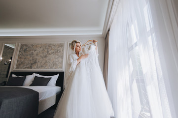 Fototapeta na wymiar Beautiful blonde bride holds a white wedding dress on a hanger and smiles in a white coat and lingerie. The morning of the bride. Wedding portrait of a cute girl in a hotel. Concept and photography.