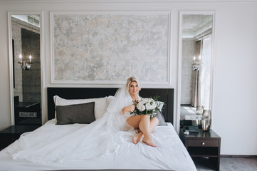 Smiling blonde bride in a white coat and lingerie, with a long veil sits on a sofa with a bouquet....