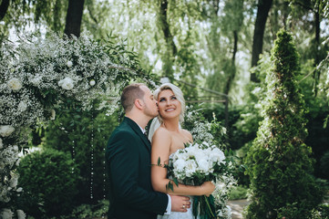 Beautiful newlyweds are standing in nature in greenery and hugging. Wedding portrait of a stylish groom in a suit and a cute blonde bride in a white dress. Concept and photography.