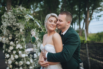 Fototapeta premium Loving newlyweds are hugging near the arch with evening decorations in the forest. Wedding portrait of a groom in love and a beautiful bride. Concept and photography.