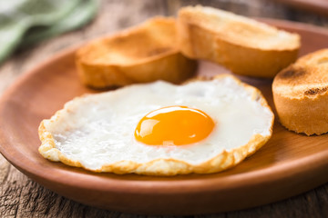One fresh fried egg sunny side up with toasted baguette slices on the side served on wooden plate (Selective Focus, Focus on the front of the egg yolk)