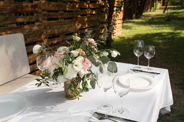 Wedding table setting decorated with fresh flowers in a brass vase. Wedding floristry. Banquet table for guests outdoors with a view of green nature. Bouquet with roses, eustoma and eucalyptus leaves