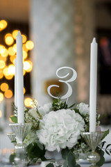 Wedding table setting is decorated with fresh flowers and white candles. Wedding floristry. Bouquet with roses, hydrangea and eustoma. On the background blur are burning garlands with light bulbs