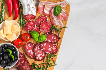 Cutting board with french salami, smoked sausage, bacon, cheese, olives and herbs on white marble background