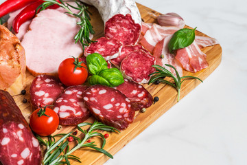 Salami, sliced ham, sausage, bacon. Meat antipasto platter with vegetables and herbs on white marble table