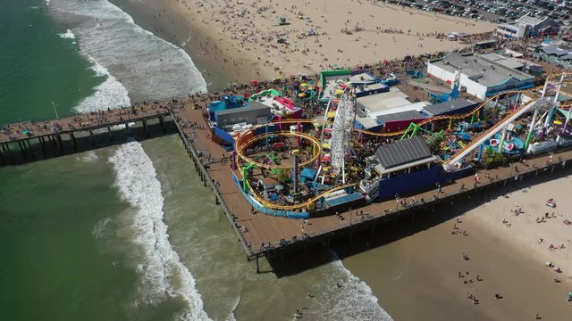 Aerial: Looking Down At The Colorful Santa Monica Pier, Tourists, And Aquamarine Waves Crashing On The Beach  - Santa Monica, California