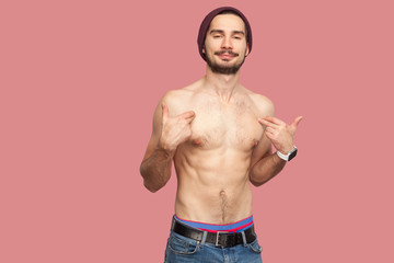 This is me! Portrait of proud handsome naked shirtless young bearded man standing, pointing and showing himself and looking at camera with flirty face. indoor studio shot, isolated on pink background.