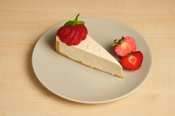 A plate with a strawberry decorated cheesecake stands on a light wooden table.