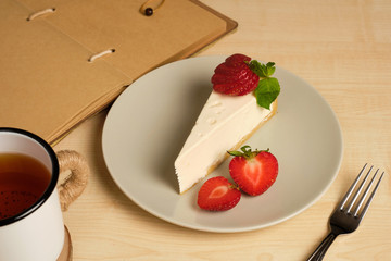A plate with cheesecake and strawberries on a light table stands along with a cup of tea and a notebook