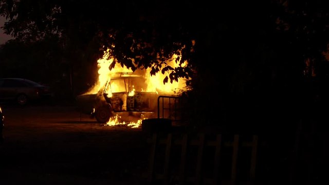 Burning car in a residential area of city. Theme of riots criminal arson of a car near house. Automobile engulfed in flames of fire. Trees close to burning sedan, slow motion.