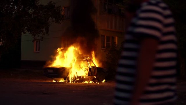 Car on fire near a residential building explodes and continues to burn. Theme of riots criminal arson of a car near house. Trees close to burning sedan. A man walks past a burning Automobile.