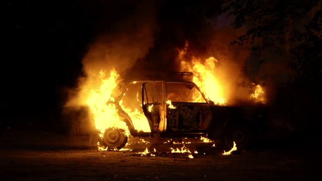 Car sedan on fire side view slow motion. Theme of criminal revenge and set fire to someone else car. Automobile engulfed in flames of fire. Trees close to burning sedan.