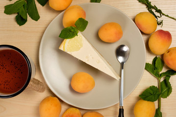 Cheesecake decorated with mint and lemon on a plate with a cup of tea is surrounded by herbs and apricots on a light wooden table