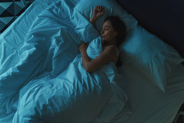 Young beautiful girl or woman sleeping alone in big bed at night, top view, blue toned