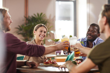 Multi-ethnic group of friends clinking glasses while sitting at table in cafe enjoying cold refreshing drinks, copy space
