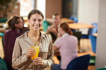 Waist up portrait of cheerful young woman looking at camera while holding refreshing drink in cafe,...